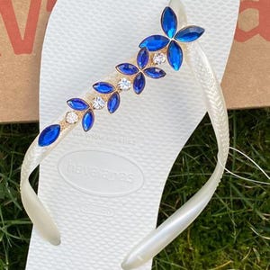 May include: White Havaianas flip-flop with a decorative strap featuring blue and clear crystal-like embellishments. The strap has a gold-coloured base. The flip-flop is on a green grassy surface.
