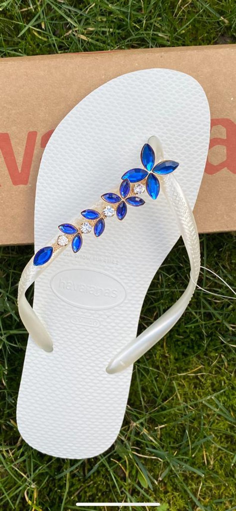 May include: White Havaianas flip-flop with a decorative strap featuring blue and clear crystal-like embellishments. The strap has a gold-coloured base. The flip-flop is on a green grassy surface.