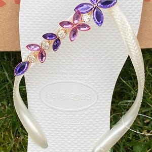 May include: White Havaianas flip-flop with a decorative floral design. The embellishment features purple, pink, and clear crystals. The flip-flop is a size EUR 41/42.