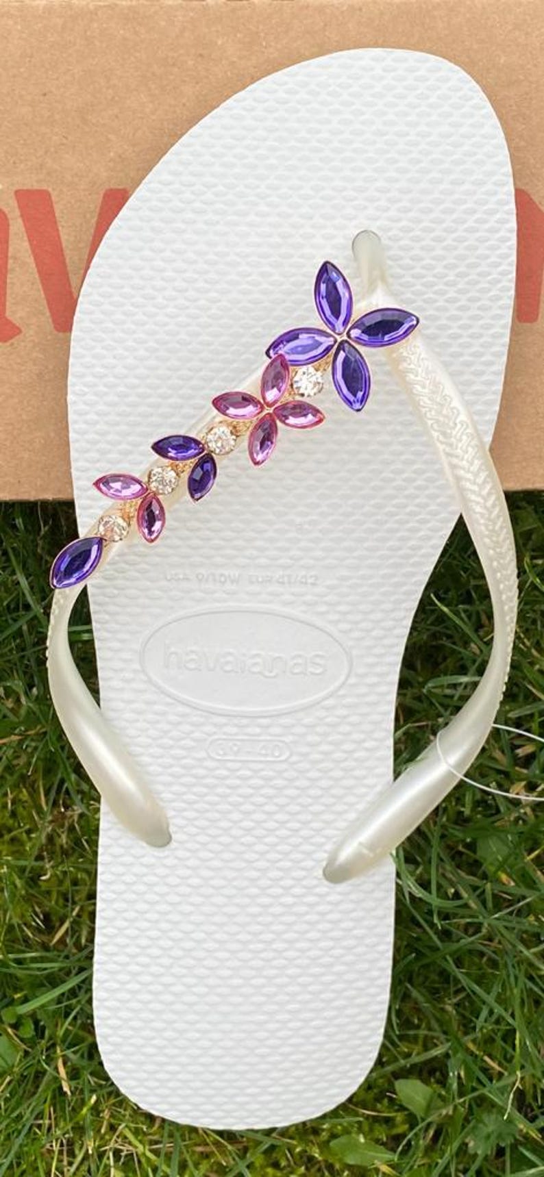 May include: White Havaianas flip-flop with a decorative floral design. The embellishment features purple, pink, and clear crystals. The flip-flop is a size EUR 41/42.