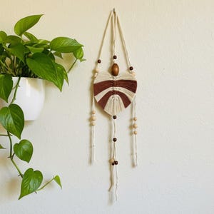 Peut inclure: Une suspension murale en macramé présentant un motif de feuille en forme de cœur, réalisé avec des cordons en coton alternant les couleurs crème et marron. L'objet est orné de perles en bois naturel de tailles variées.