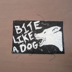 May include: Black rectangular patch with the text "BITE LIKE A DOG" in white, handwritten-style lettering. A white illustration of a snarling dog is on the right side of the patch. The patch is made of a dark denim material.