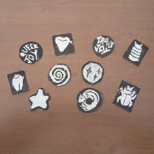 May include: A selection of black and white patches with diverse designs. Some patches display text, including "Queer Joy" and "Trans Vol". Other designs include a tooth, a spiral, a star, and an insect.