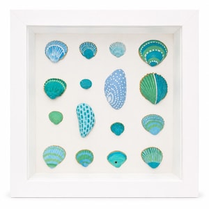 May include: A square white shadow box frame containing a collection of fifteen seashells arranged in a grid. The shells are painted in various shades of blue, teal, and green with patterns including stripes, dots, and solid colours on a white background.