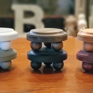 May include: Three wooden candle holders, each with a different color and finish. The first is white with a gray base, the second is dark brown with a black base, and the third is light brown with a dark brown base. Each holder has four rounded legs.