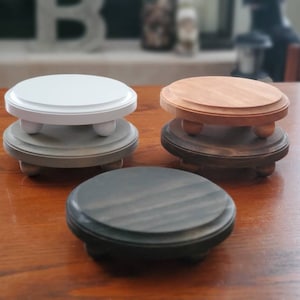 May include: Three wooden pedestal stands, one white, one natural wood, and one dark stained wood. Each stand has three legs and a round top.