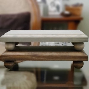 May include: A two-tiered wooden display stand. The top tier is painted a light gray, while the bottom tier is a natural wood tone. Both tiers are supported by rounded feet, creating a decorative and functional piece.