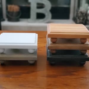 May include: Two sets of tiered wooden risers, one white, one natural wood, with a gray and black base. Each riser has four small round legs.