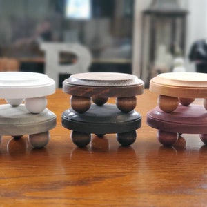 May include: Four wooden candle holders with a round top and three legs. The holders are painted in different colors: white, gray, brown, and purple.