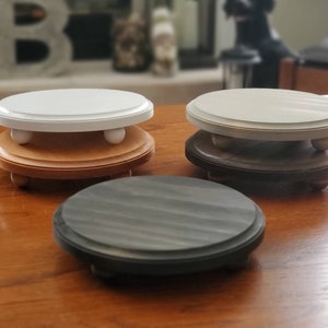 May include: Three round wooden display stands with different colored tops. The top stand is white, the middle stand is beige, and the bottom stand is gray. Each stand has three small wooden feet.