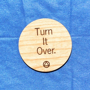 May include: A round wooden coaster with the text "Turn It Over." engraved on it. The coaster is on a blue surface.