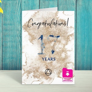 Peut inclure: Une carte blanche avec un fond aquarelle brun et du texte bleu. Le texte dit "Félicitations ! 17 ans" avec un symbole de triangle dans un cercle. La carte a un bouton rose avec une flèche de téléchargement et le texte "Téléchargement instantané".