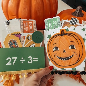 May include: A decorative scrapbook page featuring a green card with the text 27 &divide; 3=, a round green token with the number 20, orange and green admission tickets, a playing card with the number 4, and a cutout of a smiling jack-o'-lantern.
