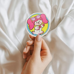 May include: A circular sticker featuring a cartoon dog with pink hair, sunglasses, and a heart-shaped necklace. The dog is white with pink accents, set against a yellow and blue background. The sticker is held against a white fabric.