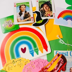 May include: A selection of Wizard of Oz themed items. Features playing cards with character portraits, a black witch's hat, a rainbow, a pair of red sequined shoes, and a green sign with the text "The Wizard of Oz".