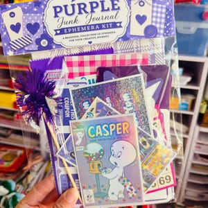 May include: A Purple Junk Journal ephemera kit in a clear plastic bag. The header reads Purple Junk Journal Ephemera Kit. Inside are various paper scraps, a Casper the Friendly Ghost comic card, and a purple tinsel wand on a wooden stick.