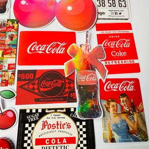 May include: A collection of Coca-Cola themed stickers and cards featuring red and white branding, vintage advertisements, a holographic bottle sticker with a bow, cherry illustrations, a bingo card, and a Postie's diet cola label with text.