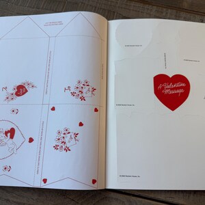 May include: Open book with Valentine's Day themed cutouts and designs. The left page features red heart and floral illustrations, with text that says "I Love You". The right page has a large red heart with "A Valentine Message" and a small "For You" label.