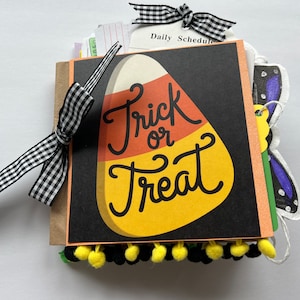 May include: A square scrapbook with a black cover featuring a large candy corn graphic and the text Trick or Treat. The book is bound with a black and white gingham ribbon bow and decorated with a row of small yellow and black pom-poms at the bottom.