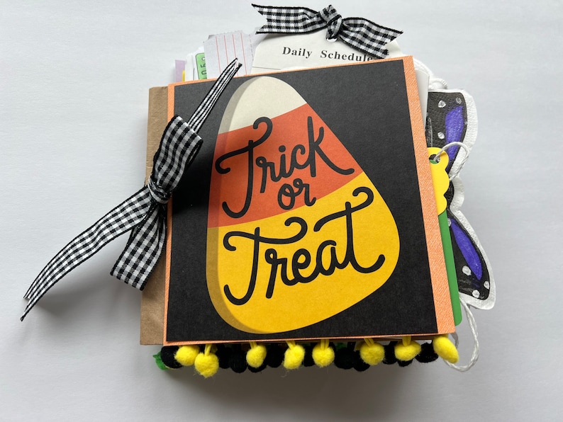 May include: A square scrapbook with a black cover featuring a large candy corn graphic and the text Trick or Treat. The book is bound with a black and white gingham ribbon bow and decorated with a row of small yellow and black pom-poms at the bottom.