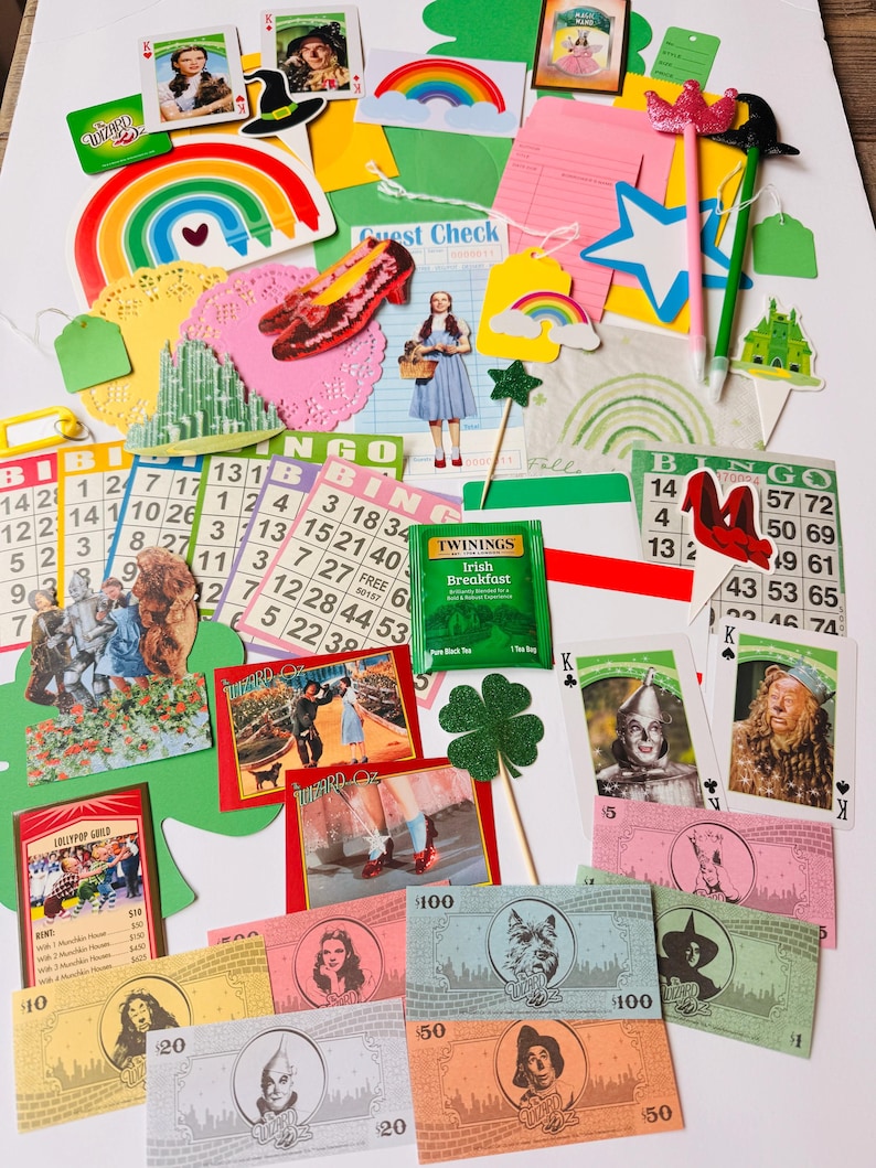 May include: A selection of Wizard of Oz themed items, such as bingo cards, playing cards, paper cutouts, and play money. The items feature characters, rainbows, and the Emerald City. A tea bag is also included.