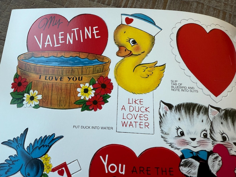 May include: A sheet of vintage Valentine's Day cutouts. Designs include a red heart with "My Valentine" text, a yellow duck in a sailor hat, and two kittens. Other elements include a wooden tub with "I Love You" text and flowers.