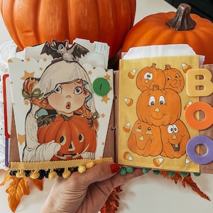 May include: A handmade scrapbook page featuring a vintage-style illustration of a child in a ghost costume holding a jack-o'-lantern, topped with a bat. The adjacent page displays a stack of cartoon pumpkins, candy corn, and the word BOO in foam letters.