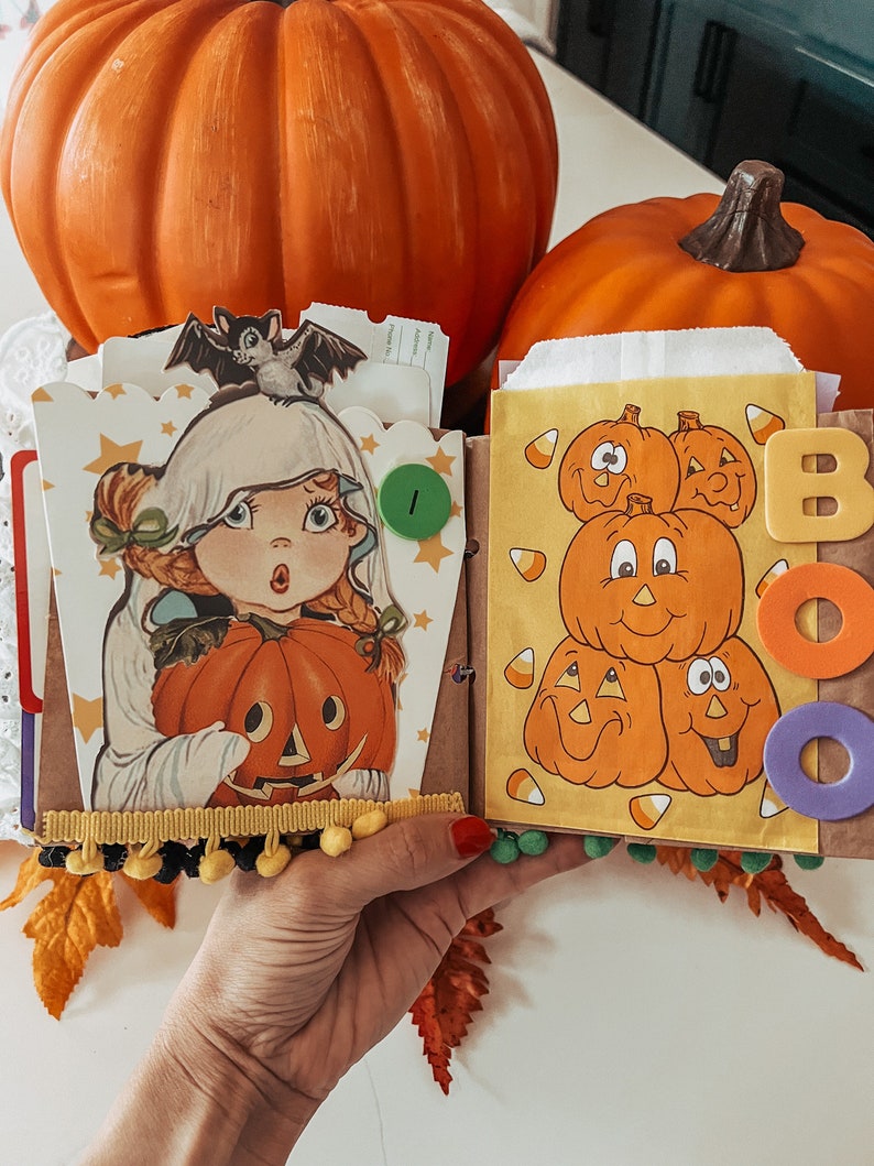 May include: A handmade scrapbook page featuring a vintage-style illustration of a child in a ghost costume holding a jack-o'-lantern, topped with a bat. The adjacent page displays a stack of cartoon pumpkins, candy corn, and the word BOO in foam letters.