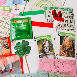 May include: A selection of Wizard of Oz themed items, including playing cards, bingo cards, and paper cutouts. A green tea bag, a green glitter shamrock, and play money are also visible. The items are arranged on a white surface.