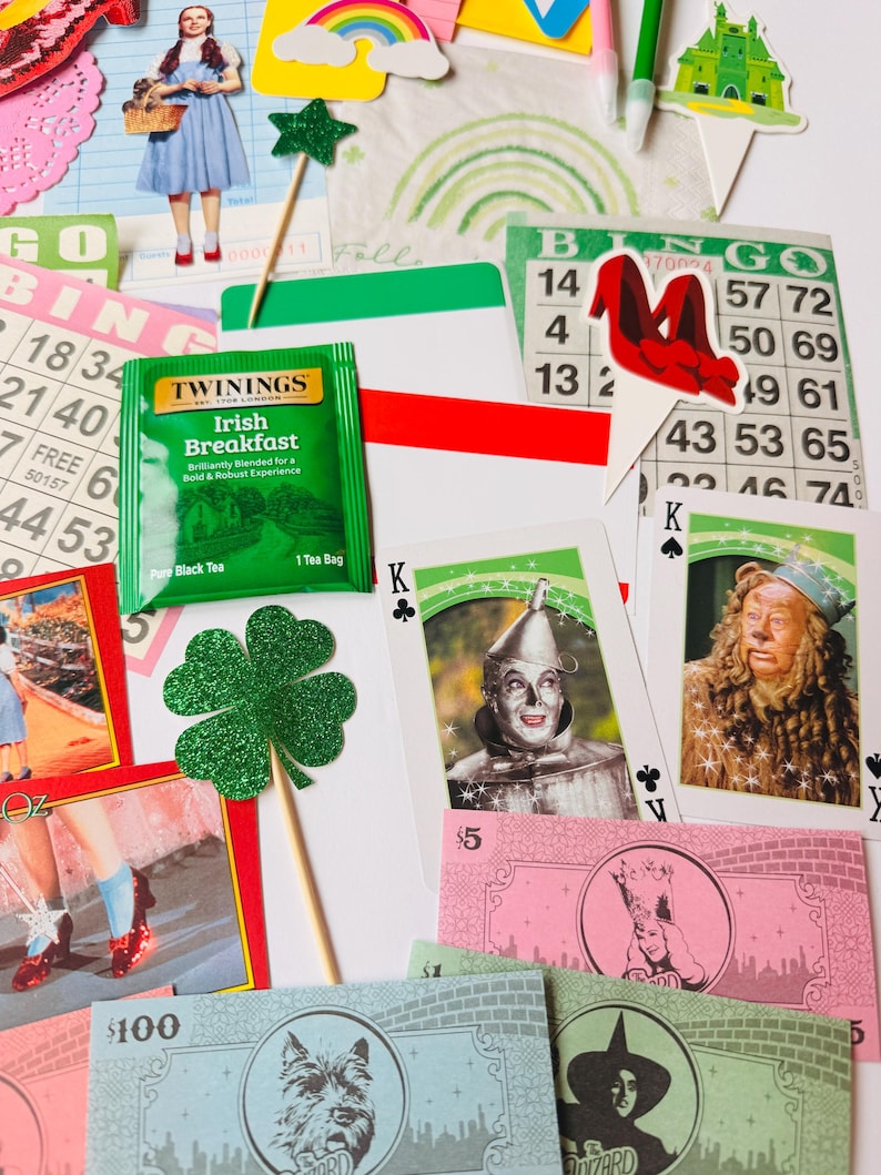 May include: A selection of Wizard of Oz themed items, including playing cards, bingo cards, and paper cutouts. A green tea bag, a green glitter shamrock, and play money are also visible. The items are arranged on a white surface.