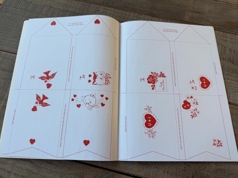 May include: Open book with pages featuring red illustrations on a white background. Designs include birds, cats, teddy bears, roses, and hearts. The text "I + U" is visible on some of the hearts. The pages are divided into sections with dotted lines.