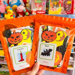 Puede incluir: Dos bolsas de plástico naranja que contienen recortes de papel y tarjetas con temática de Halloween. Las tarjetas visibles muestran ilustraciones de una vela roja, un murciélago negro y calabazas naranjas, con etiquetas de texto como Candle, Bat y Cross.