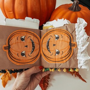 May include: A brown paper craft book featuring two orange pumpkin cutouts with vintage-style faces on the front cover. The book is decorated with white eyelet lace trim on the right edge and small yellow and green pom-poms along the bottom edge.