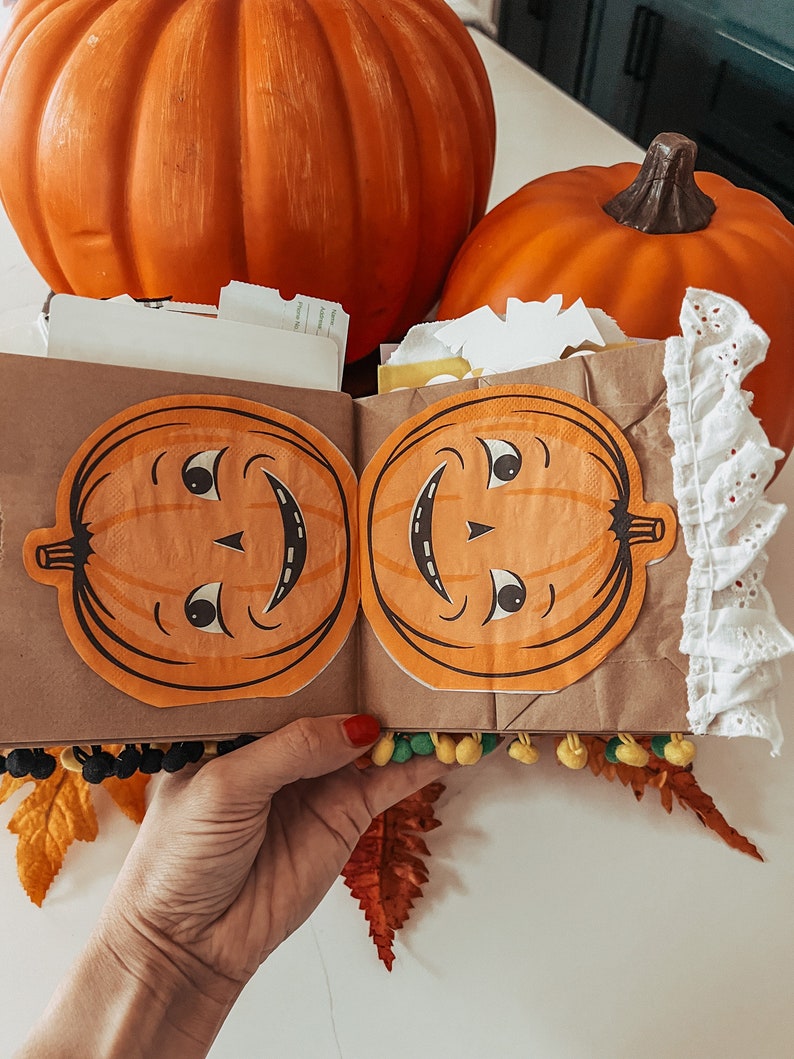 May include: A brown paper craft book featuring two orange pumpkin cutouts with vintage-style faces on the front cover. The book is decorated with white eyelet lace trim on the right edge and small yellow and green pom-poms along the bottom edge.