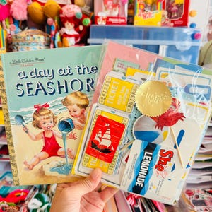 Puede incluir: Un libro Little Golden Book titulado A Day at the Seashore con una ilustración vintage de niños jugando en la arena, junto a una bolsa de plástico transparente con diversos artículos de papel, incluyendo boletos amarillos, una pegatina de barco roja y una etiqueta de limonada.