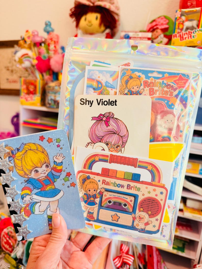 K&ouml;nnte beinhalten: Eine Sammlung von Schreibwaren im Rainbow Brite-Design, bestehend aus einem kleinen Notizbuch mit Charakterillustration, einem holografischen Beutel mit Aufklebern sowie Karten mit Shy Violet und einer bunten Kassettenrekorder-Grafik.