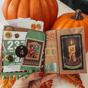 May include: A brown paper craft book featuring Halloween-themed ephemera, including a coat check ticket numbered 237, a small green envelope with a witch illustration, a black circular tag with the number 4, and a Knight of Candles tarot card.