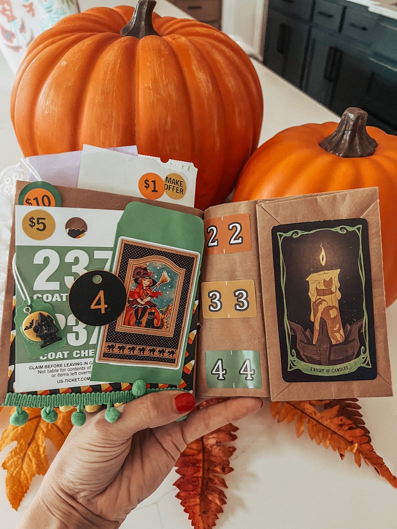 May include: A brown paper craft book featuring Halloween-themed ephemera, including a coat check ticket numbered 237, a small green envelope with a witch illustration, a black circular tag with the number 4, and a Knight of Candles tarot card.
