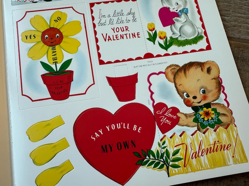 May include: A sheet of vintage Valentine's Day cutouts. Designs include a smiling flower with "Yes," "No," and "Maybe" petals, a bunny holding a heart, and a teddy bear with a heart that says "I love you."