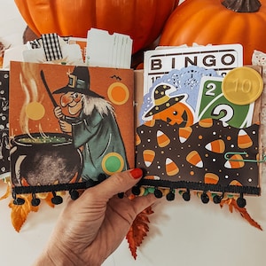 May include: A Halloween-themed scrapbook page featuring a vintage-style illustration of a witch stirring a black cauldron. The page includes a pocket filled with a Bingo card, a green number two playing card, and a pumpkin cutout with a hat.
