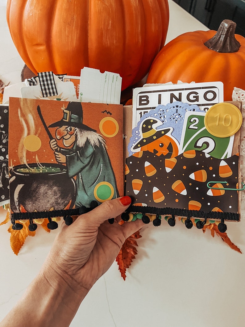 May include: A Halloween-themed scrapbook page featuring a vintage-style illustration of a witch stirring a black cauldron. The page includes a pocket filled with a Bingo card, a green number two playing card, and a pumpkin cutout with a hat.