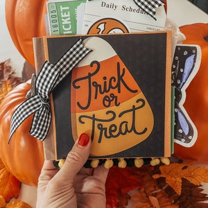 May include: A square scrapbook with a black cover featuring a large candy corn graphic and the text Trick or Treat. The book is decorated with a black and white gingham ribbon bow and yellow pom-pom trim, containing various paper inserts and tickets.