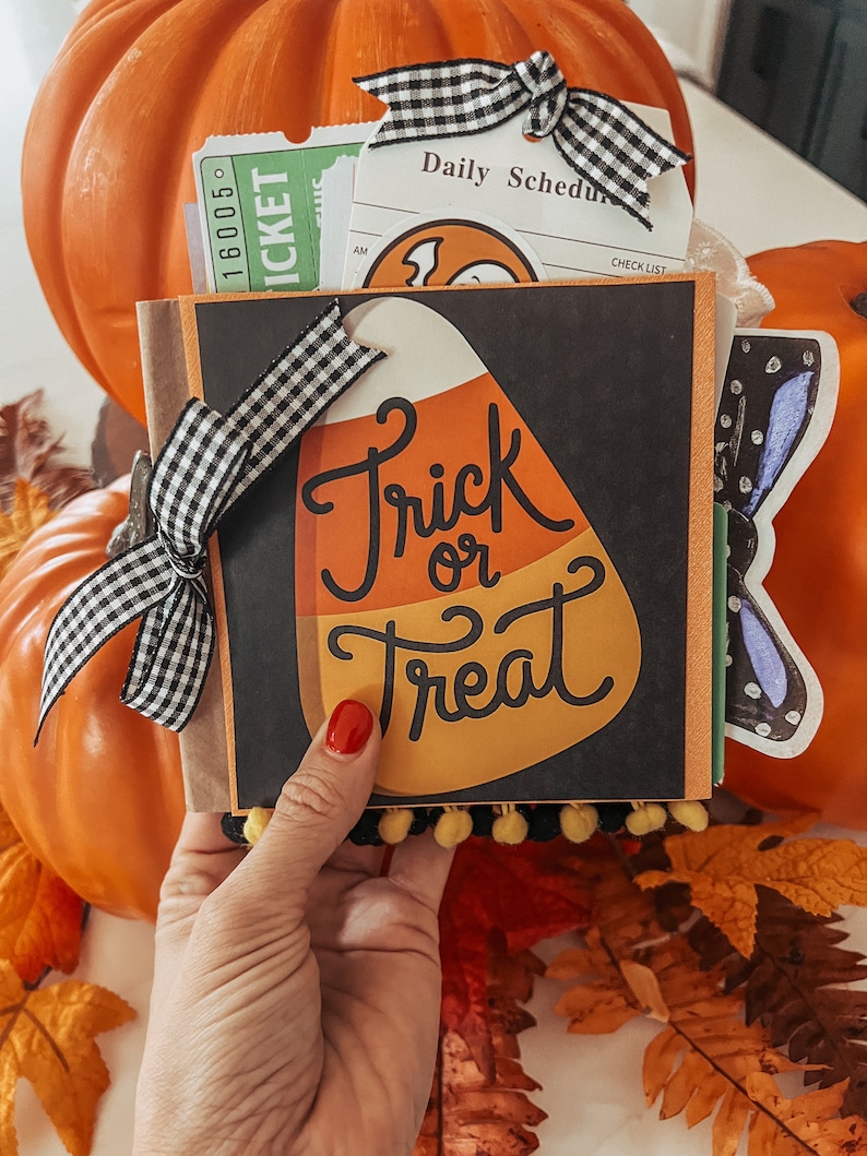 May include: A square scrapbook with a black cover featuring a large candy corn graphic and the text Trick or Treat. The book is decorated with a black and white gingham ribbon bow and yellow pom-pom trim, containing various paper inserts and tickets.