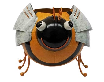 May include: A whimsical metal bee-shaped planter with a smiling face, black and orange body, and silver-colored wings. The planter has three pairs of curved legs and is designed to hold small plants or flowers. The bee's body is approximately 8 inches in diameter.