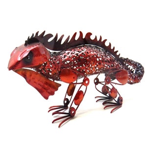 May include: A red metal iguana sculpture with a textured body and spiky dorsal crest. The iguana has detailed legs and a head with visible eyes. The sculpture is made of metal and is a decorative item.