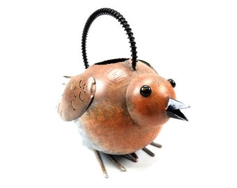 May include: A metal bird-shaped planter with a brown body, black eyes, and a black handle. The bird has a small, open beak and is perched on three small legs. The planter is designed for outdoor use and adds a whimsical touch to any garden.