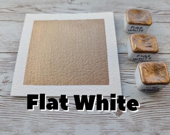 May include: A square of textured, flat white paint on a white paper, with three small, square paint containers. The containers have gold-colored tops and the words "flat white" written on them. The text "Flat White" is displayed below the paper.