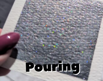 May include: A square of textured, silver material with iridescent flecks, set on a white background. The word "Pouring" is printed in bold black letters across the bottom of the image. The material has a rough, glittery appearance.