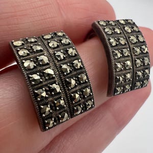 Può includere: Un paio di gemelli vintage color argento. Ogni gemello presenta un design rettangolare con file di piccole pietre scintillanti. Le pietre sono ravvicinate, creando una superficie strutturata. I gemelli sono curvi e hanno un aspetto classico ed elegante.