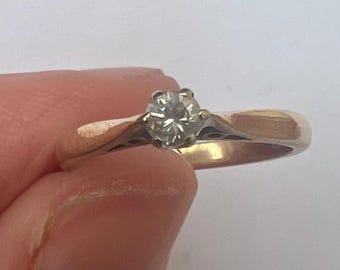 May include: A gold ring featuring a single, round, clear diamond. The ring has a plain band and a four-prong setting. The ring is held by a person's fingers.